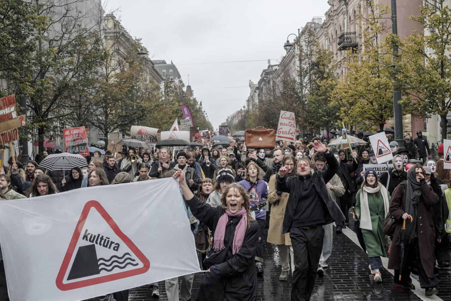 Crowd of people march in the rain in protest. A woman at the front holds a banner that shows a red triangular road sign with the word "kultura" sliding off a cliff into waves