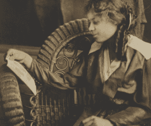A black-and-white photograph of a young girl sitting on a wicker chair. The young girl is wearing a dark color, loose fitting dress with a light color neckline. She reads from a piece of paper in her left hand. The photograph has a brown mat frame and backing.